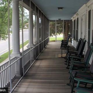 A long screened porch with wooden flooring and green rocking chairs lined up along the side.