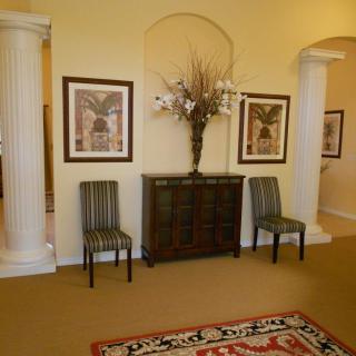 A well-appointed living room with elegant decor, featuring a central vase on a cabinet, framed paintings, two chairs, and decorative columns.