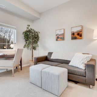 A cozy living room featuring a gray sofa, two ottomans, a dining area by the window, and decorative wall art.