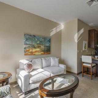 A cozy living room with a white sofa and glass coffee tables, featuring a landscape painting on the wall and flooded with natural light.