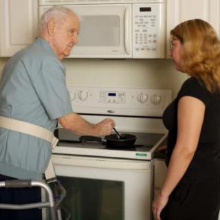 An elderly man using a walker receives assistance from a caregiver in the kitchen.