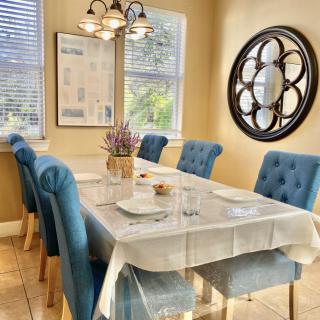 A modern dining room with a neatly set table and blue upholstered chairs, featuring a decorative wall mirror and ample natural light.
