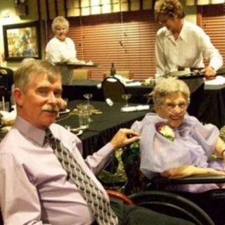 A family gathering at a restaurant, featuring an elderly woman and a man in a tie, with waitstaff serving in the background.
