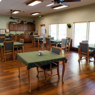 A spacious dining room with several tables and chairs, featuring a wooden floor and a kitchen area in the background.