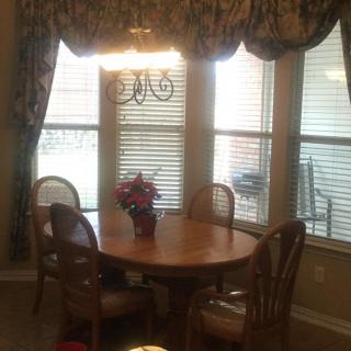 A cozy dining room with a wooden table and chairs, adorned by a chandelier and surrounded by large windows with curtains.
