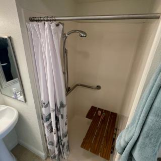 A small bathroom featuring a shower with a curtain and bath mat, adjacent to a sink and mirror.