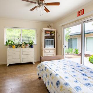 A bright bedroom with a ceiling fan, large windows, and sliding doors opening to the outside. It features a dresser with plants and floral bedding.