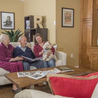 An elderly couple sits on a sofa with a younger woman holding a small dog, enjoying a cozy living room setting.