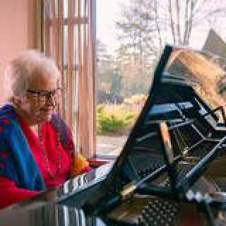 An elderly woman playing the piano by a sunlit window.