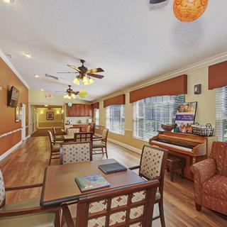 A cozy interior dining space featuring tables, chairs, and a piano, with large windows and warm lighting.