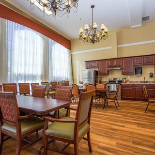 A spacious dining area with a kitchen, featuring a wooden floor, elegant chandeliers, and large windows with curtains.