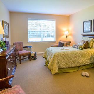 A cozy bedroom featuring a neatly made bed, a lamp, chairs, and a window providing natural light.