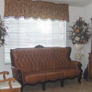 A traditional living room with a leather sofa, large window with blinds and curtains, and decorative floral arrangements.