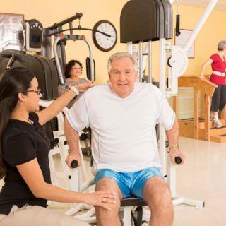 An elderly man receives guidance from a trainer while using exercise equipment in a gym.