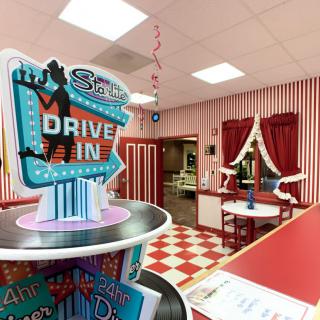 A retro-themed diner with red and white decor, featuring a 'Drive-In' sign and a classic checkerboard floor.