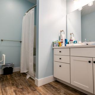 A modern bathroom with light blue walls, featuring a shower, vanity with a large mirror, and wooden flooring.