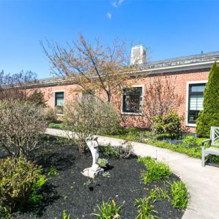 A charming garden with a winding pathway and a bench, set against a brick building under a clear blue sky.