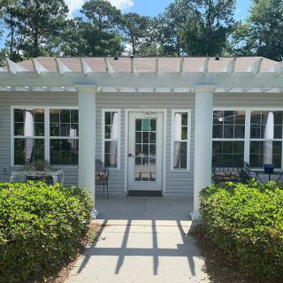 A white house entrance with a pergola and surrounding greenery under a clear blue sky.