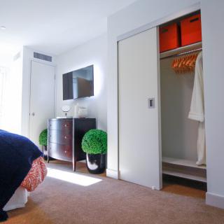A bright and modern bedroom with a television, dresser, and an open closet displaying robes. The room is decorated with potted plants.