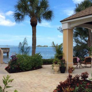 A beautiful waterfront patio with a tropical vibe, featuring a palm tree, outdoor seating, and lush landscaping.