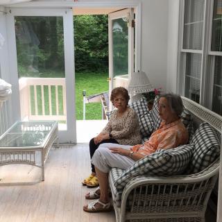 Two people sitting on a porch, engaged in conversation with a view of greenery outside.