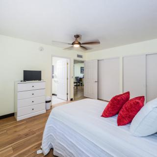 A cozy bedroom featuring a bed with red pillows, white linens, a white dresser, and a ceiling fan. The room is decorated with hardwood floors and neutral walls.