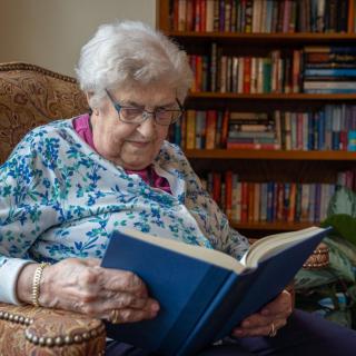 An elderly woman reading a book while sitting in an armchair, surrounded by bookshelves.