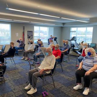 A group of seniors participating in an exercise class while seated in chairs, all wearing masks inside a community center.