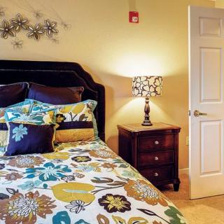 A cozy bedroom featuring a bed with floral-patterned bedding and decorative pillows, next to a nightstand with a lamp.