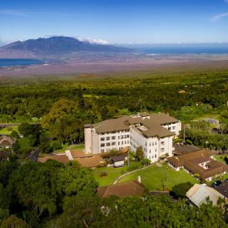 Aerial view of a building set amidst lush greenery with a mountainous coastline in the background.