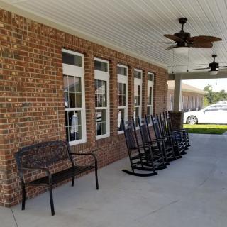 A porch with multiple rocking chairs, a bench, and a ceiling fan along a brick wall.