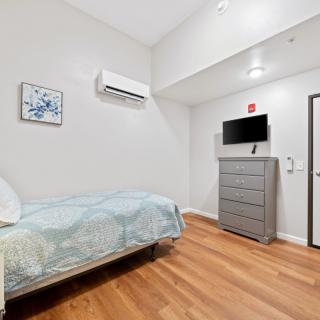 A minimalist bedroom with wooden flooring, featuring a single bed, dresser, mounted television, and air conditioner.