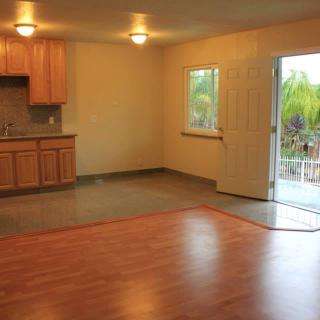 An empty room featuring a small kitchen area with wooden cabinets, an open door leading outside, and a window allowing natural light.