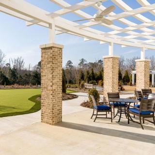 An outdoor patio area with a pergola, featuring tables and chairs, surrounded by a garden landscape.