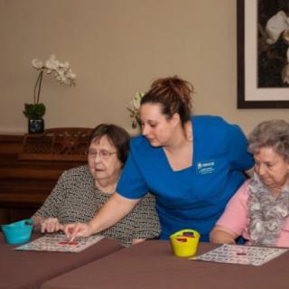 A caregiver assists elderly women during a bingo game in a nursing home setting.