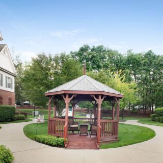 A charming wooden gazebo surrounded by a lush green lawn and pathway, set against a backdrop of trees and a residential building.