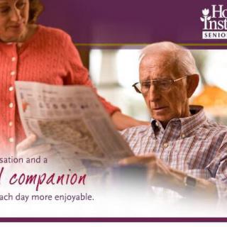 An elderly man reading a newspaper with a caregiver providing companionship at home.