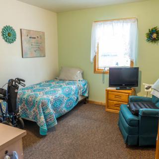 A cozy bedroom with a single bed, wheelchair, television, and armchair by the window.