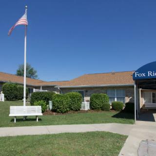 A building with an American flag, surrounded by grass and bushes, featuring a bench and an awning at the entrance.