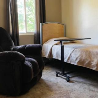A cozy bedroom with a single bed, recliner, and adjustable side table beside a window.