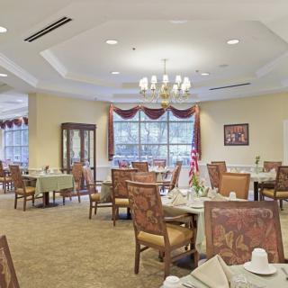 A well-lit dining room featuring elegantly set tables and chairs with a chandelier and large windows adorned with curtains.