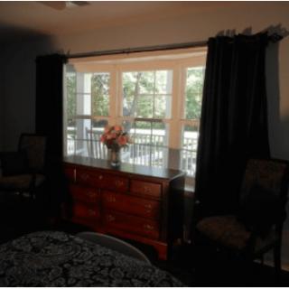 A cozy bedroom featuring a dresser with flowers, flanked by chairs and framed by dark curtains and a window.