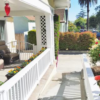 A sunny view of a porch with seating, surrounded by a garden and pathway in a residential area.