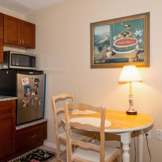 A cozy kitchen corner with a small dining table and chairs, illuminated by a table lamp. A painting hangs on the wall above the warm wooden cabinetry.