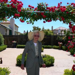 A person in a suit and wide-brimmed hat stands in a garden under a floral archway.