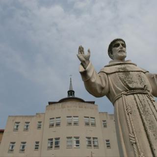 A statue of a religious figure stands prominently in front of a building against a cloudy sky.