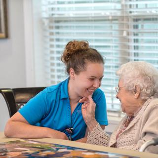 A caregiver and a senior woman share a warm moment of conversation indoors.