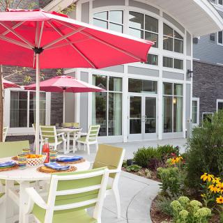A cozy outdoor patio area with tables, chairs, and red umbrellas surrounded by a garden.