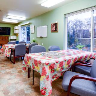 A cozy dining area with tables covered in floral tablecloths, surrounded by chairs and illuminated by bright ceiling lights.