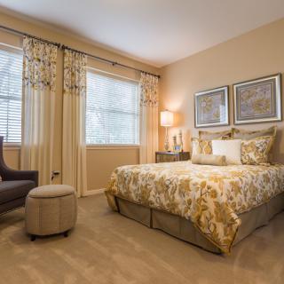 A well-decorated bedroom featuring a bed with patterned bedding, matching curtains, a lamp, and a cozy chair.